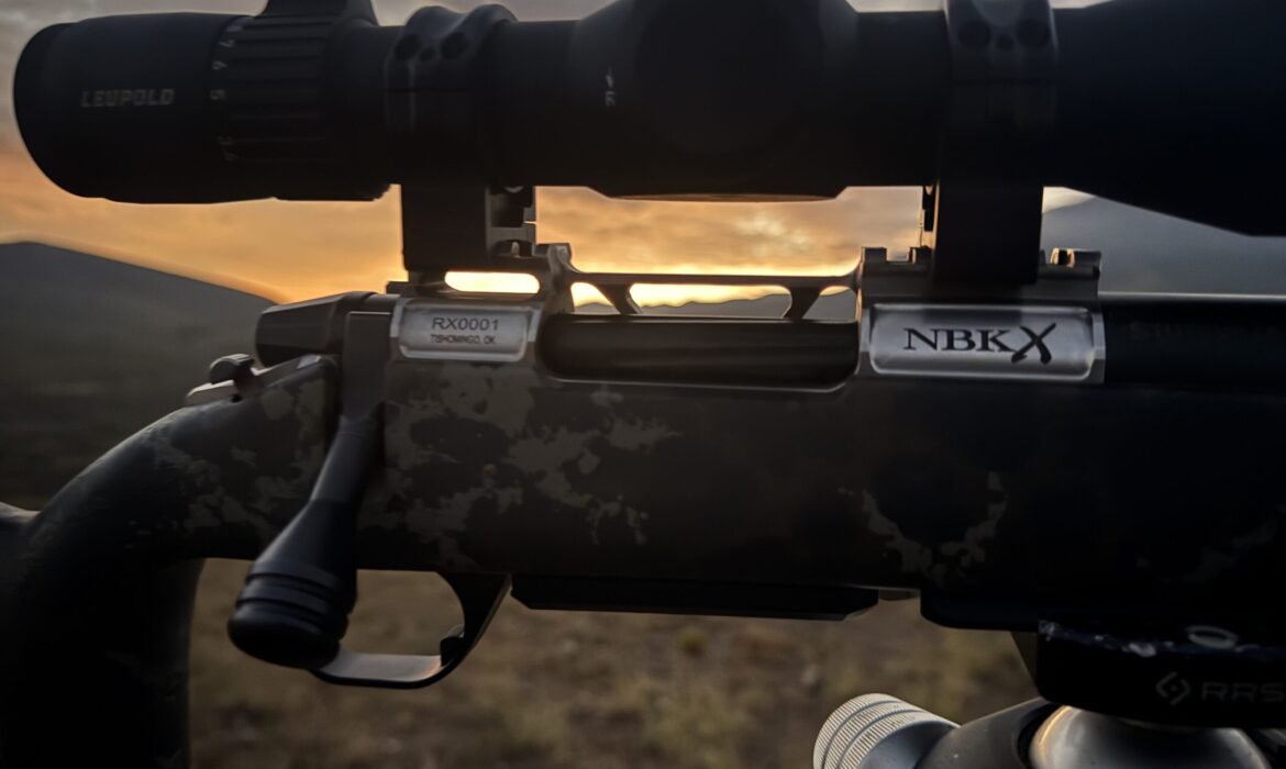 Close-up of a rifle scope mounted on a camouflaged rifle with Leupold branding, sunrise in the background.