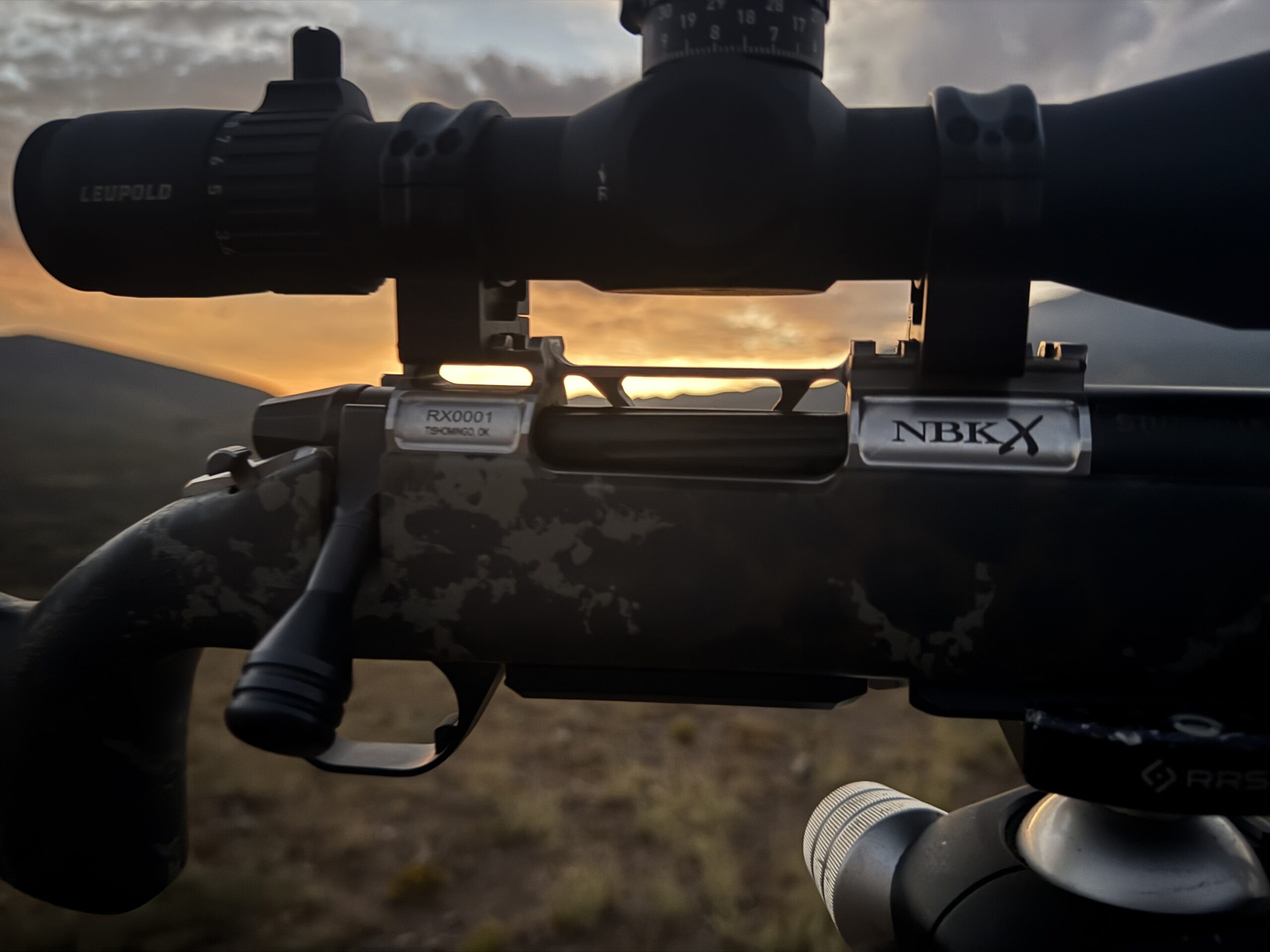 Close-up of a rifle scope mounted on a camouflaged rifle with Leupold branding, sunrise in the background.