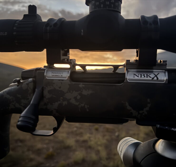 Close-up of a rifle scope mounted on a camouflaged rifle with Leupold branding, sunrise in the background.