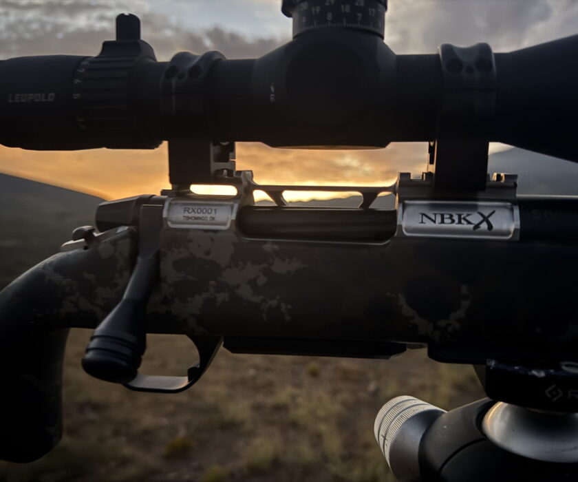 Close-up of a rifle scope mounted on a camouflaged rifle with Leupold branding, sunrise in the background.